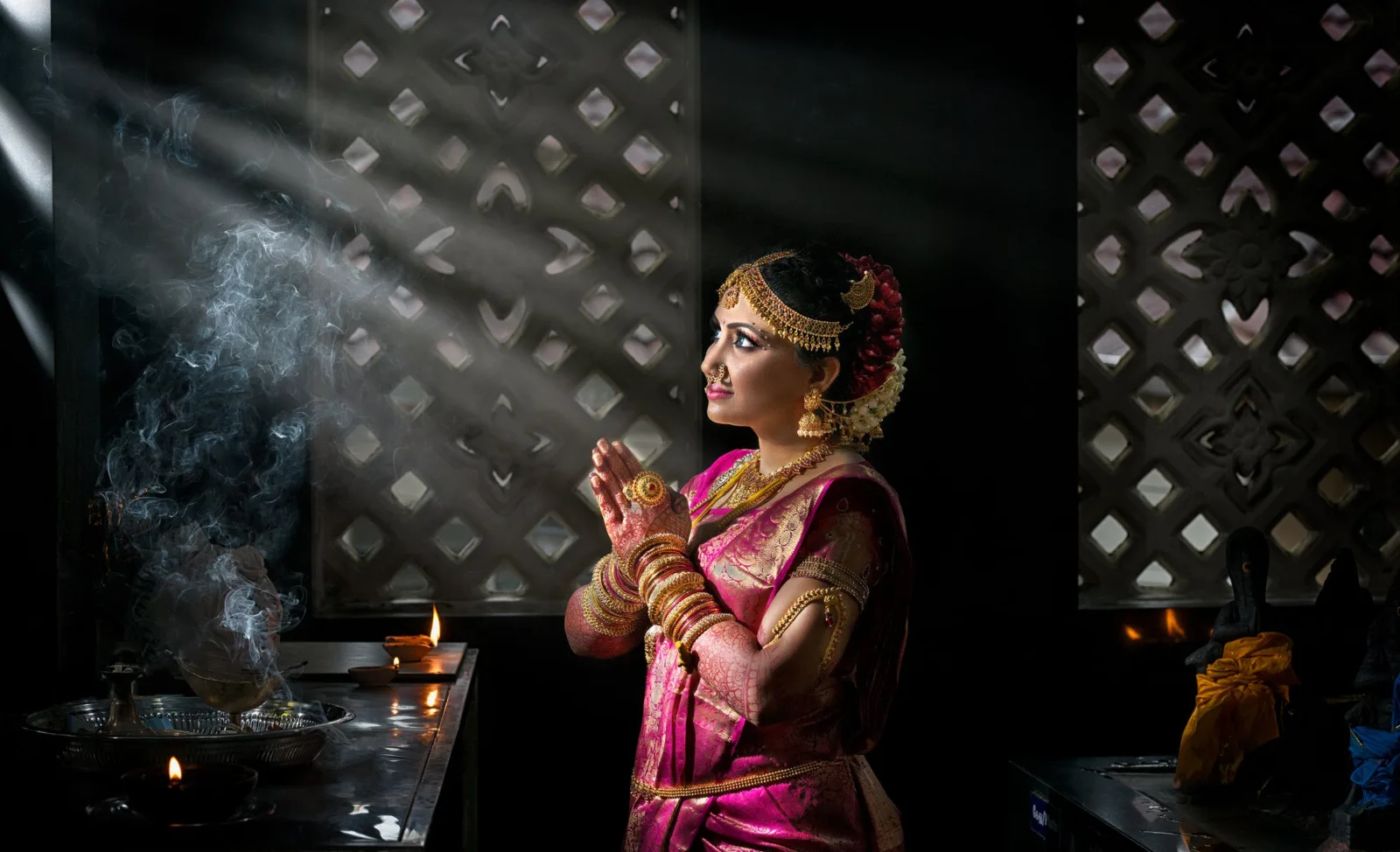 Bride in a pink silk saree praying with incense smoke and window light rays