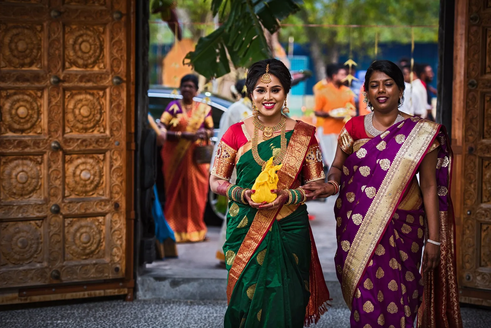Indian bride in a green and red silk saree walking through carved temple doors with her bridesmaid